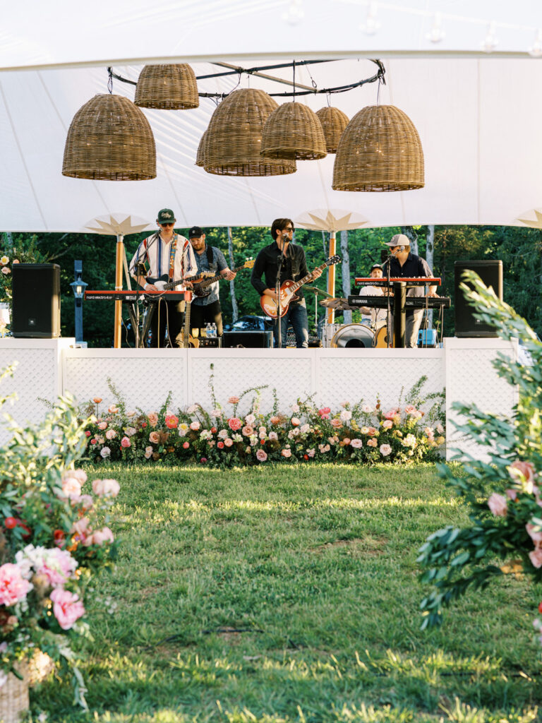 Flowers arranged to create a lush, vibrant border that complements the white lattice stage facade. The selection of flowers, ranging from roses to peonies and other seasonal blooms, adds texture and depth. Installation features a mix of heights to create a natural, garden-like feel. Sailcloth private evet with basket pendant lighting and spring florals. Design by Rosemary & Finch Floral Design. 