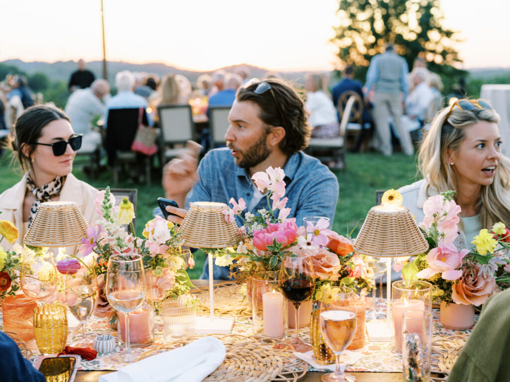 Elegantly set outdoor table with a picturesque view of the downtown Nashville skyline. Tables adorned with floral tablecloths featuring a colorful and intricate pattern. Each table has floral centerpieces with a mix of pink, peach, and yellow flowers, adding a vibrant touch to the setting. Floral design for garden inspired birthday party. Peony inspired floral design with bright spring florals. Design by Rosemary & Finch Floral Design in Nashville, TN. 