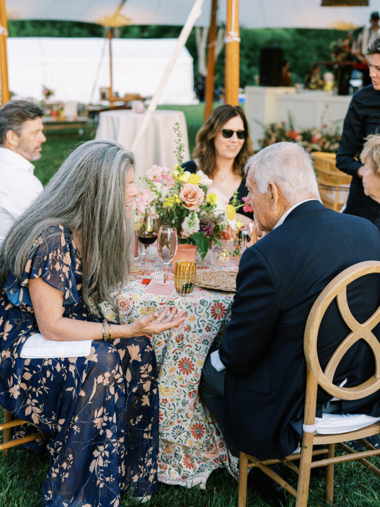 Elegantly set outdoor table with a picturesque view of the downtown Nashville skyline. Tables adorned with floral tablecloths featuring a colorful and intricate pattern. Each table has floral centerpieces with a mix of pink, peach, and yellow flowers, adding a vibrant touch to the setting. Floral design for garden inspired birthday party. Peony inspired floral design with bright spring florals. Design by Rosemary & Finch Floral Design in Nashville, TN. 