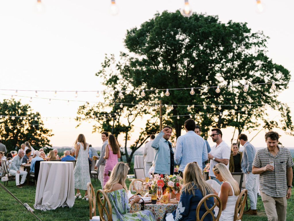 Elegantly set outdoor table with a picturesque view of the downtown Nashville skyline. Tables adorned with floral tablecloths featuring a colorful and intricate pattern. Each table has floral centerpieces with a mix of pink, peach, and yellow flowers, adding a vibrant touch to the setting. Floral design for garden inspired birthday party. Peony inspired floral design with bright spring florals. Design by Rosemary & Finch Floral Design in Nashville, TN. 