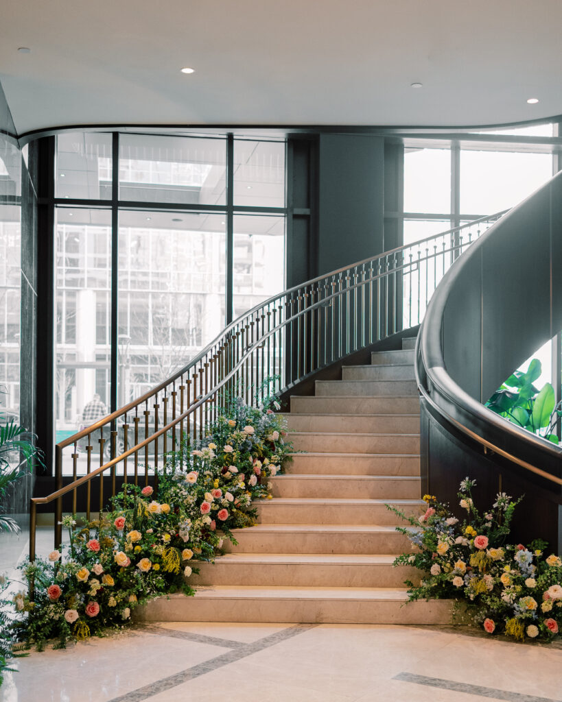 Garden-inspired floral installation on winding staircase. Flowers consisting of soft and vibrant hues, including shades of pink, peach, yellow, cream, and touches of burgundy, adding depth and interest. Winter wedding in downtown Nashville. Unique ceremony entrance for winter wedding. Design by Rosemary & Finch Floral Design. 