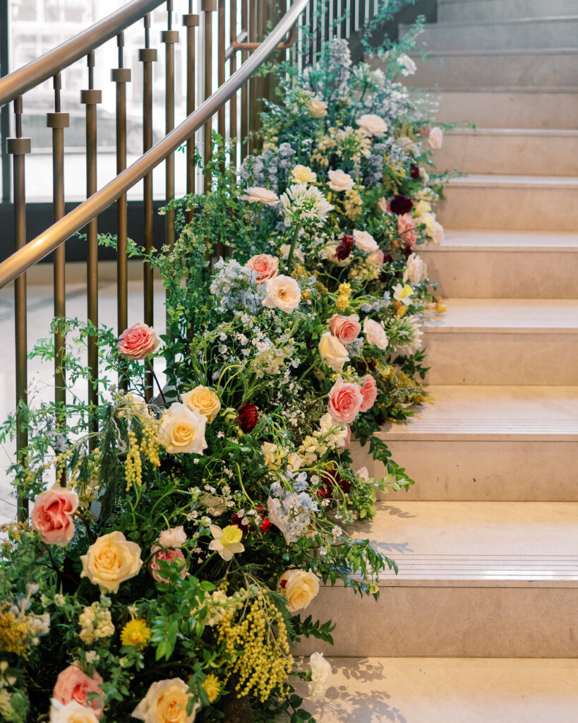 Garden-inspired floral installation on winding staircase. Flowers consisting of soft and vibrant hues, including shades of pink, peach, yellow, cream, and touches of burgundy, adding depth and interest. Winter wedding in downtown Nashville. Unique ceremony entrance for winter wedding. Design by Rosemary & Finch Floral Design. 