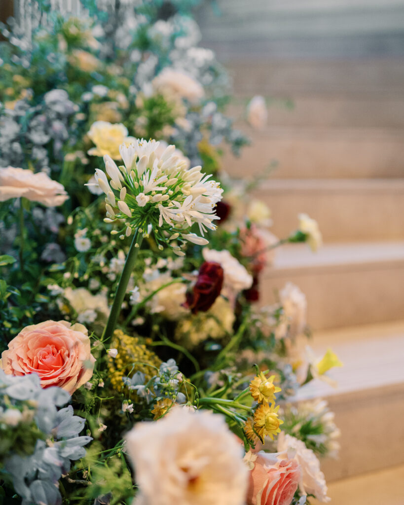 Garden-inspired floral installation on winding staircase. Flowers consisting of soft and vibrant hues, including shades of pink, peach, yellow, cream, and touches of burgundy, adding depth and interest. Winter wedding in downtown Nashville. Unique ceremony entrance for winter wedding. Design by Rosemary & Finch Floral Design. 