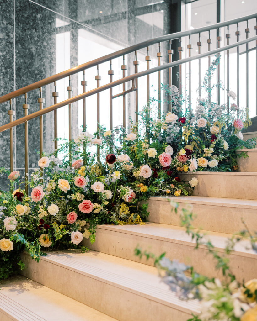 Garden-inspired floral installation on winding staircase. Flowers consisting of soft and vibrant hues, including shades of pink, peach, yellow, cream, and touches of burgundy, adding depth and interest. Winter wedding in downtown Nashville. Unique ceremony entrance for winter wedding. Design by Rosemary & Finch Floral Design. 