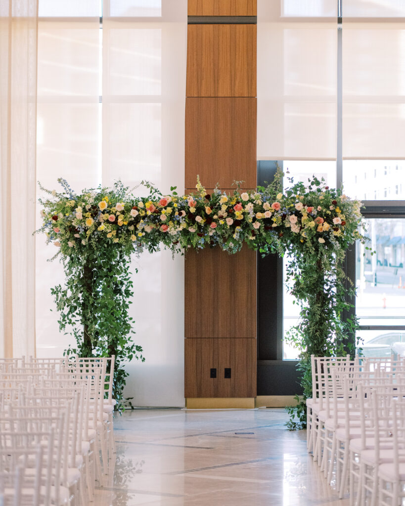 Winter wedding ceremony chuppah wrapped densely in cascading greenery, giving the structure a natural, garden-like appearance. The flowers consisting of soft and vibrant hues, including shades of pink, peach, yellow, cream, and touches of burgundy, adding depth and interest. Winter wedding in downtown Nashville. Design by Rosemary & Finch Floral Design. 