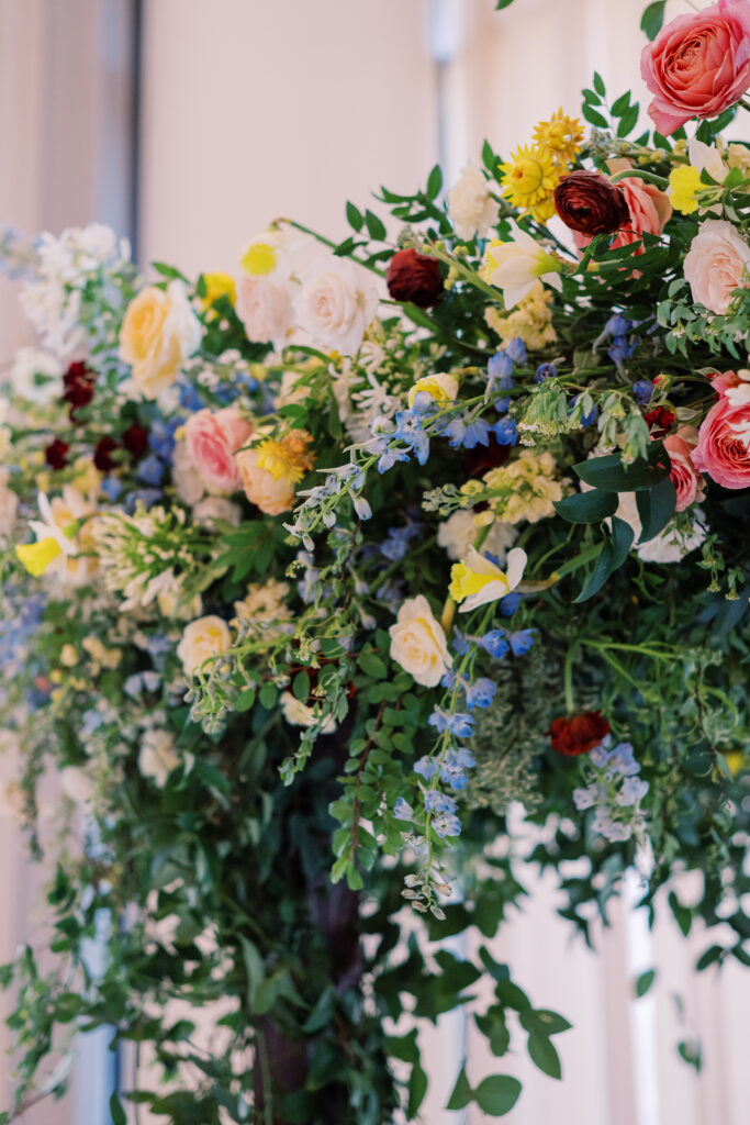 Winter wedding ceremony chuppah wrapped densely in cascading greenery, giving the structure a natural, garden-like appearance. The flowers consisting of soft and vibrant hues, including shades of pink, peach, yellow, cream, and touches of burgundy, adding depth and interest. Winter wedding in downtown Nashville. Design by Rosemary & Finch Floral Design. 