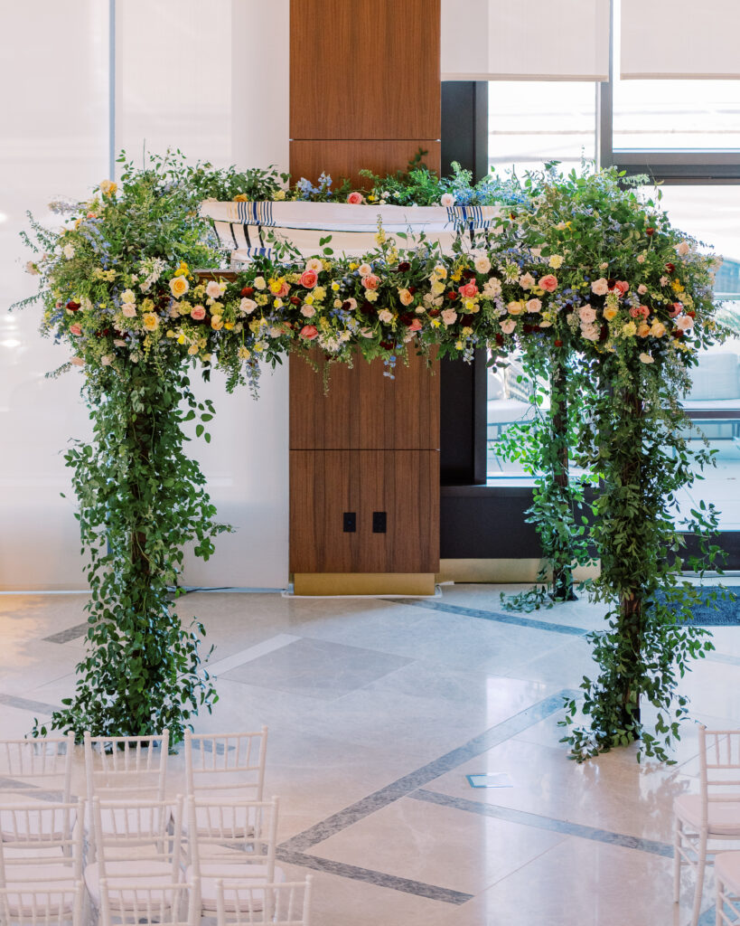 Winter wedding ceremony chuppah wrapped densely in cascading greenery, giving the structure a natural, garden-like appearance. The flowers consisting of soft and vibrant hues, including shades of pink, peach, yellow, cream, and touches of burgundy, adding depth and interest. Winter wedding in downtown Nashville. Design by Rosemary & Finch Floral Design. 