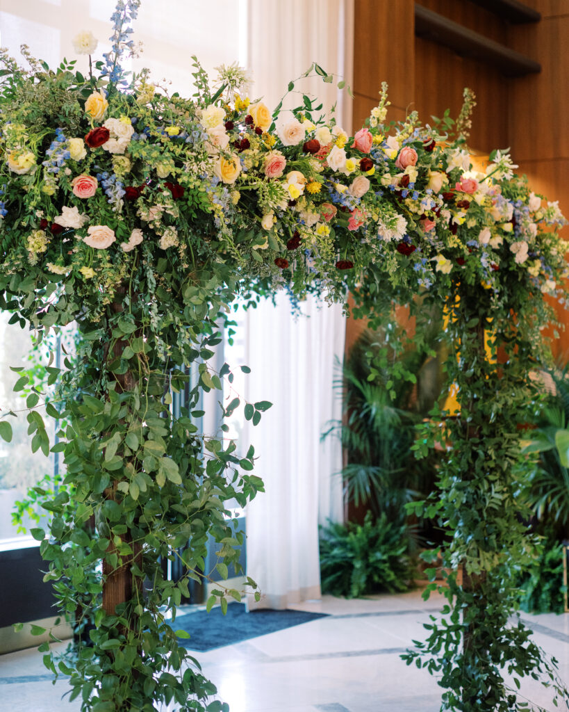 Winter wedding ceremony chuppah wrapped densely in cascading greenery, giving the structure a natural, garden-like appearance. The flowers consisting of soft and vibrant hues, including shades of pink, peach, yellow, cream, and touches of burgundy, adding depth and interest. Winter wedding in downtown Nashville. Design by Rosemary & Finch Floral Design. 