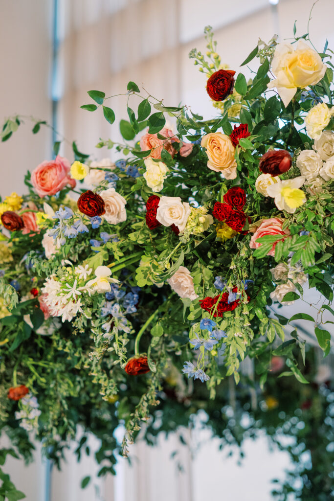 Winter wedding ceremony chuppah wrapped densely in cascading greenery, giving the structure a natural, garden-like appearance. The flowers consisting of soft and vibrant hues, including shades of pink, peach, yellow, cream, and touches of burgundy, adding depth and interest. Winter wedding in downtown Nashville. Design by Rosemary & Finch Floral Design. 