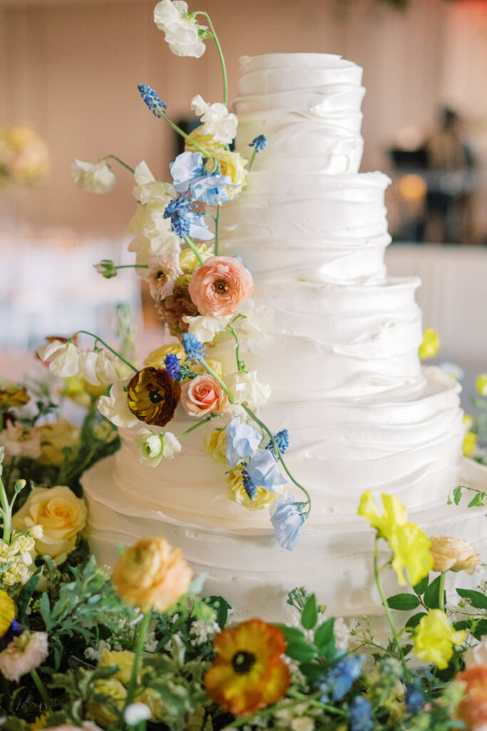 Lush floral cake meadows create memorable moment for wedding reception cake cutting. Elegant winter wedding color palette in reds, yellows, cream, and soft hints of blue. Lush garden-inspired floral installations. Design by Rosemary & Finch Floral Design. 