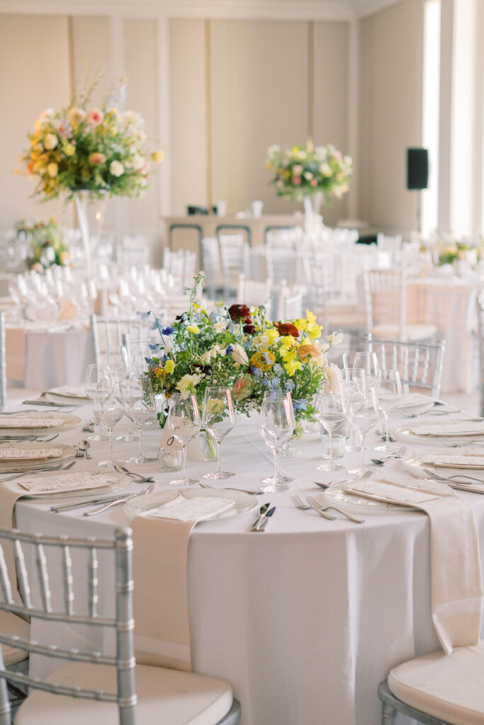 Elegant wedding reception tables feature low lush floral centerpieces and elevated centerpieces in glass vases. The centerpiece is a beautiful arrangement of mixed flowers in soft pastel colors, including yellows, whites, and some darker tones like red and burgundy. Winter wedding reception with floral emphasis and clean lines. Taupe candles in clear glass holders create romantic candlelight. Design by Rosemary & Finch Floral Design. 