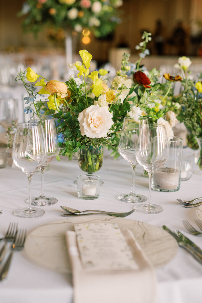 Elegant wedding reception tables feature low lush floral centerpieces and elevated centerpieces in glass vases. The centerpiece is a beautiful arrangement of mixed flowers in soft pastel colors, including yellows, whites, and some darker tones like red and burgundy. Winter wedding reception with floral emphasis and clean lines. Taupe candles in clear glass holders create romantic candlelight. Design by Rosemary & Finch Floral Design. 