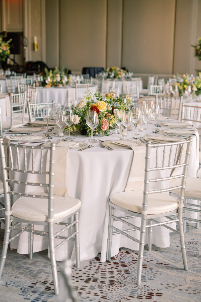 Elegant wedding reception tables feature low lush floral centerpieces and elevated centerpieces in glass vases. The centerpiece is a beautiful arrangement of mixed flowers in soft pastel colors, including yellows, whites, and some darker tones like red and burgundy. Winter wedding reception with floral emphasis and clean lines. Taupe candles in clear glass holders create romantic candlelight. Design by Rosemary & Finch Floral Design. 