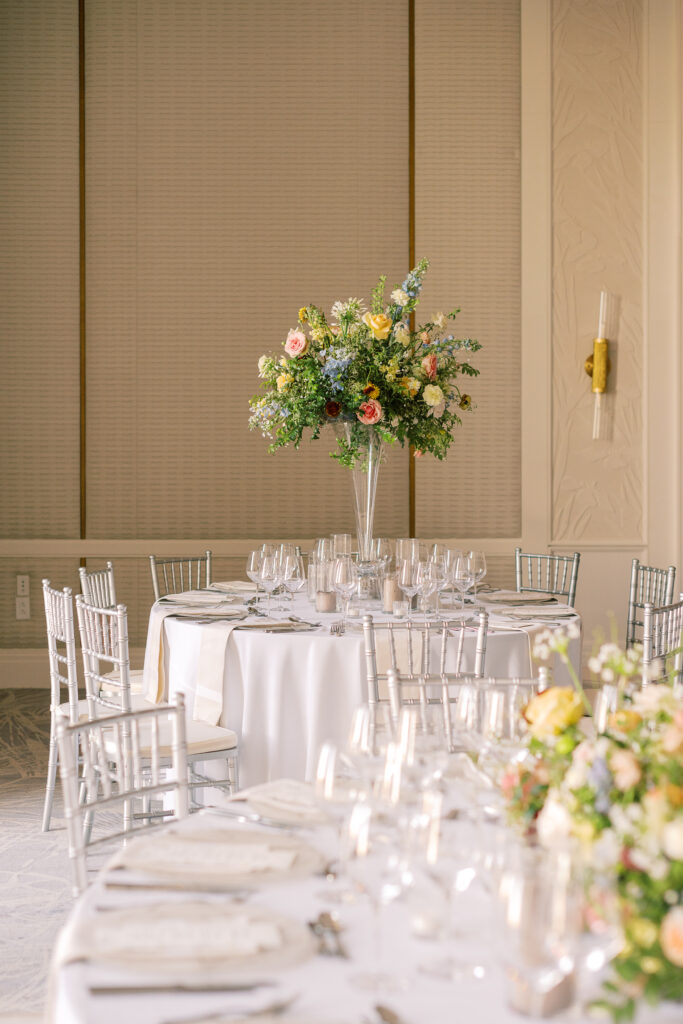 Elegant wedding reception tables feature low lush floral centerpieces and elevated centerpieces in glass vases. The centerpiece is a beautiful arrangement of mixed flowers in soft pastel colors, including yellows, whites, and some darker tones like red and burgundy. Winter wedding reception with floral emphasis and clean lines. Taupe candles in clear glass holders create romantic candlelight. Design by Rosemary & Finch Floral Design. 