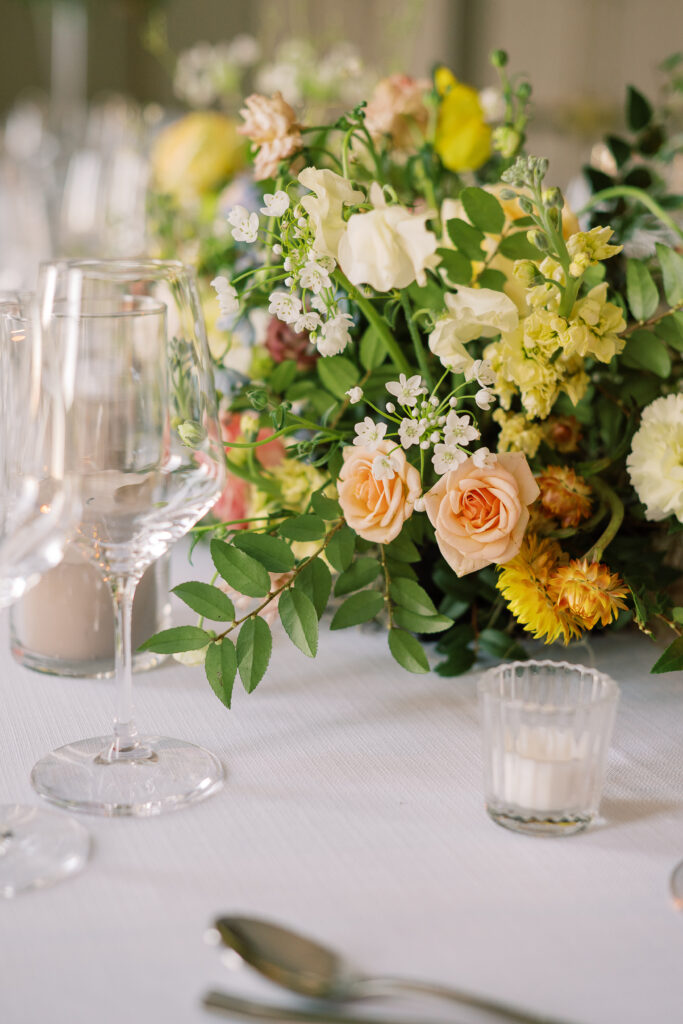 Elegant wedding reception tables feature low lush floral centerpieces and elevated centerpieces in glass vases. The centerpiece is a beautiful arrangement of mixed flowers in soft pastel colors, including yellows, whites, and some darker tones like red and burgundy. Winter wedding reception with floral emphasis and clean lines. Taupe candles in clear glass holders create romantic candlelight. Design by Rosemary & Finch Floral Design. 