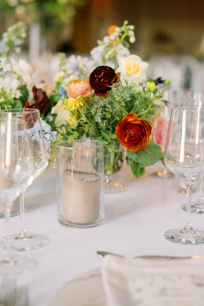 Elegant wedding reception tables feature low lush floral centerpieces and elevated centerpieces in glass vases. The centerpiece is a beautiful arrangement of mixed flowers in soft pastel colors, including yellows, whites, and some darker tones like red and burgundy. Winter wedding reception with floral emphasis and clean lines. Taupe candles in clear glass holders create romantic candlelight. Design by Rosemary & Finch Floral Design. 