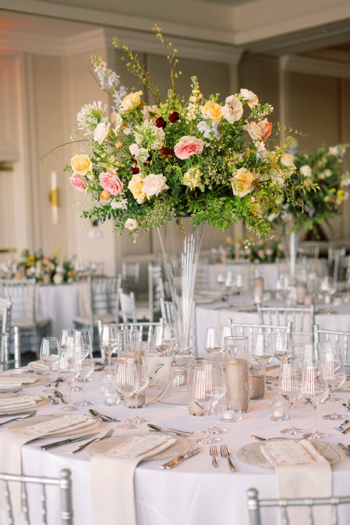 Elegant wedding reception tables feature low lush floral centerpieces and elevated centerpieces in glass vases. The centerpiece is a beautiful arrangement of mixed flowers in soft pastel colors, including yellows, whites, and some darker tones like red and burgundy. Winter wedding reception with floral emphasis and clean lines. Taupe candles in clear glass holders create romantic candlelight. Design by Rosemary & Finch Floral Design. 