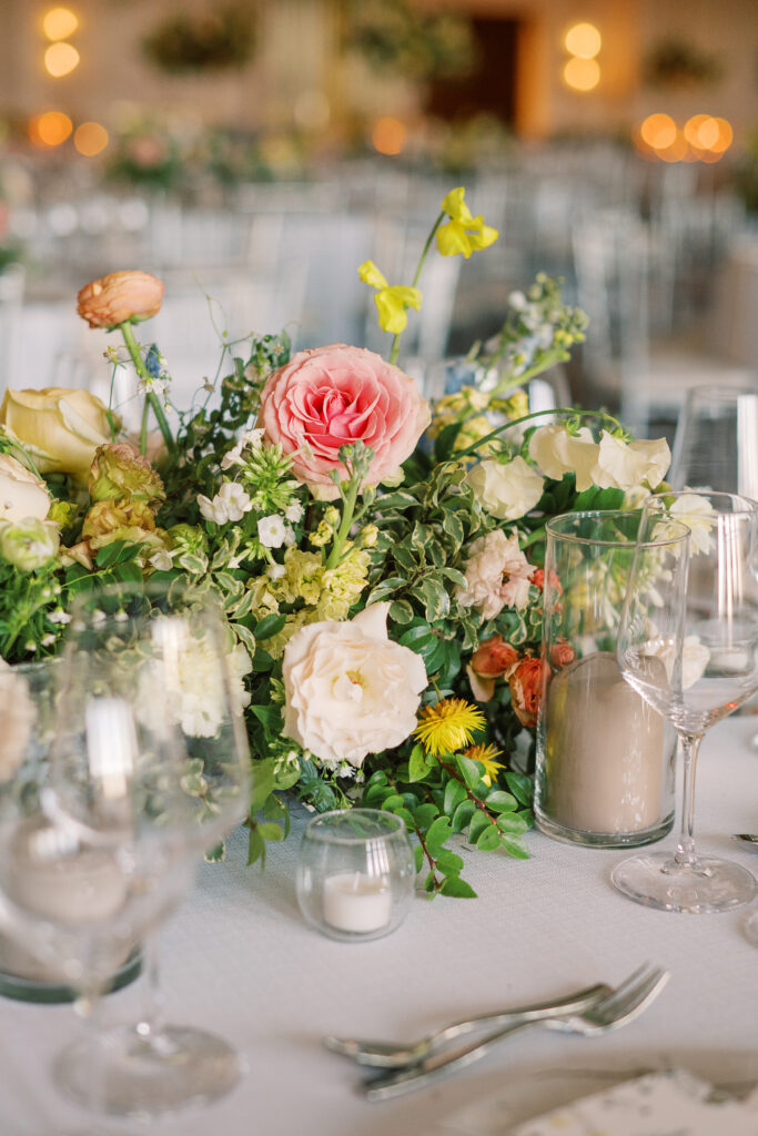 Elegant wedding reception tables feature low lush floral centerpieces and elevated centerpieces in glass vases. The centerpiece is a beautiful arrangement of mixed flowers in soft pastel colors, including yellows, whites, and some darker tones like red and burgundy. Winter wedding reception with floral emphasis and clean lines. Taupe candles in clear glass holders create romantic candlelight. Design by Rosemary & Finch Floral Design. 