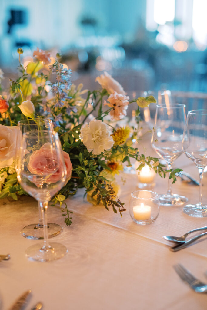 Elegant wedding reception tables feature low lush floral centerpieces and elevated centerpieces in glass vases. The centerpiece is a beautiful arrangement of mixed flowers in soft pastel colors, including yellows, whites, and some darker tones like red and burgundy. Winter wedding reception with floral emphasis and clean lines. Taupe candles in clear glass holders create romantic candlelight. Design by Rosemary & Finch Floral Design. 