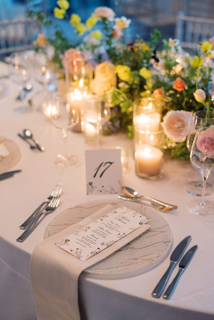 Elegant wedding reception tables feature low lush floral centerpieces and elevated centerpieces in glass vases. The centerpiece is a beautiful arrangement of mixed flowers in soft pastel colors, including yellows, whites, and some darker tones like red and burgundy. Winter wedding reception with floral emphasis and clean lines. Taupe candles in clear glass holders create romantic candlelight. Design by Rosemary & Finch Floral Design. 