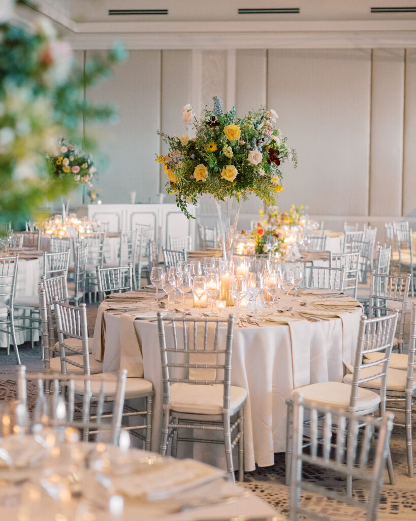 Elegant wedding reception tables feature low lush floral centerpieces and elevated centerpieces in glass vases. The centerpiece is a beautiful arrangement of mixed flowers in soft pastel colors, including yellows, whites, and some darker tones like red and burgundy. Winter wedding reception with floral emphasis and clean lines. Taupe candles in clear glass holders create romantic candlelight. Design by Rosemary & Finch Floral Design. 