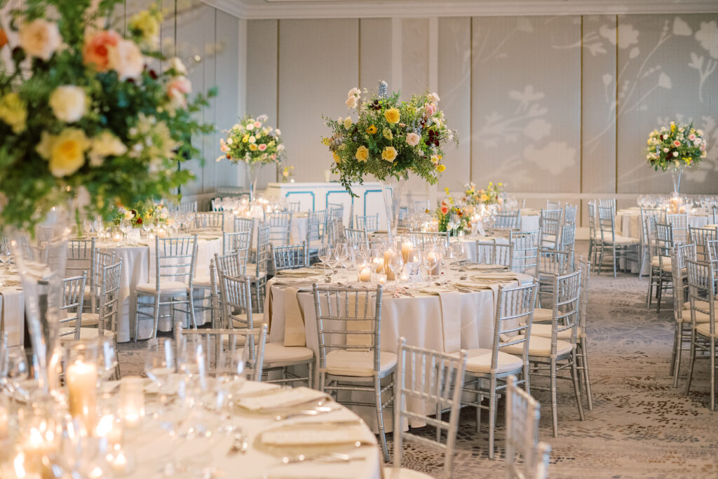 Elegant wedding reception tables feature low lush floral centerpieces and elevated centerpieces in glass vases. The centerpiece is a beautiful arrangement of mixed flowers in soft pastel colors, including yellows, whites, and some darker tones like red and burgundy. Winter wedding reception with floral emphasis and clean lines. Taupe candles in clear glass holders create romantic candlelight. Design by Rosemary & Finch Floral Design. 