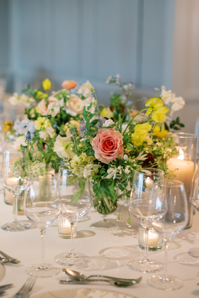 Elegant wedding reception tables feature low lush floral centerpieces and elevated centerpieces in glass vases. The centerpiece is a beautiful arrangement of mixed flowers in soft pastel colors, including yellows, whites, and some darker tones like red and burgundy. Winter wedding reception with floral emphasis and clean lines. Taupe candles in clear glass holders create romantic candlelight. Design by Rosemary & Finch Floral Design. 