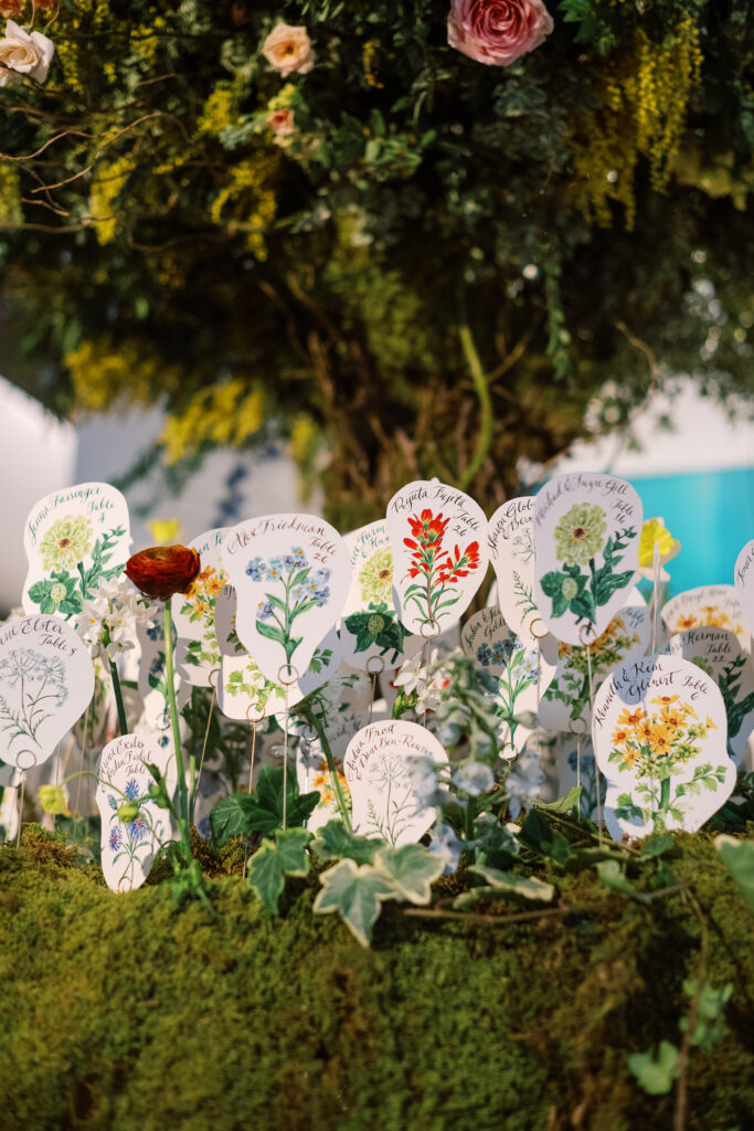 Tree escort display with hand illustrated escort cards in flower meadows. Mossy floral meadows hold escort cards for winter wedding reception. Unique wedding color palette consisting of reds, butter yellows, soft blue, and rich burgundy. Design by Rosemary & Finch Floral Design.