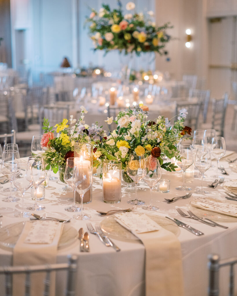 Elegant wedding reception tables feature low lush floral centerpieces and elevated centerpieces in glass vases. The centerpiece is a beautiful arrangement of mixed flowers in soft pastel colors, including yellows, whites, and some darker tones like red and burgundy. Winter wedding reception with floral emphasis and clean lines. Taupe candles in clear glass holders create romantic candlelight. Design by Rosemary & Finch Floral Design. 