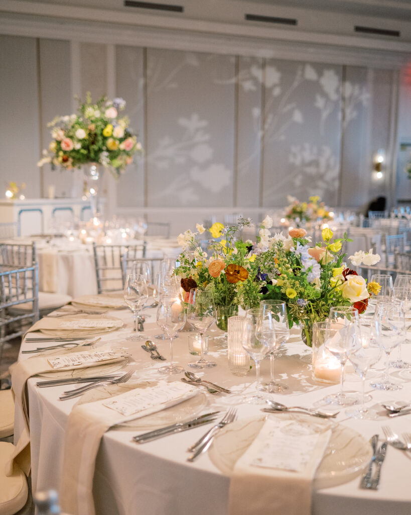 Elegant wedding reception tables feature low lush floral centerpieces and elevated centerpieces in glass vases. The centerpiece is a beautiful arrangement of mixed flowers in soft pastel colors, including yellows, whites, and some darker tones like red and burgundy. Winter wedding reception with floral emphasis and clean lines. Taupe candles in clear glass holders create romantic candlelight. Design by Rosemary & Finch Floral Design. 