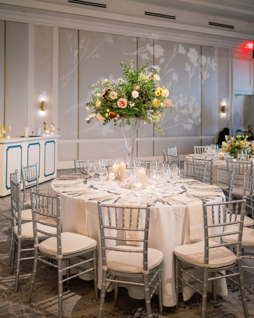 Elegant wedding reception tables feature low lush floral centerpieces and elevated centerpieces in glass vases. The centerpiece is a beautiful arrangement of mixed flowers in soft pastel colors, including yellows, whites, and some darker tones like red and burgundy. Winter wedding reception with floral emphasis and clean lines. Taupe candles in clear glass holders create romantic candlelight. Design by Rosemary & Finch Floral Design. 