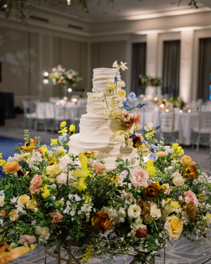 Lush floral cake meadows create memorable moment for wedding reception cake cutting. Elegant winter wedding color palette in reds, yellows, cream, and soft hints of blue. Lush garden-inspired floral installations. Design by Rosemary & Finch Floral Design. 