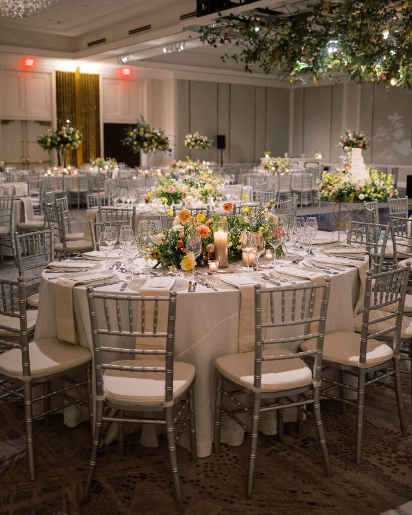 Elegant wedding reception tables feature low lush floral centerpieces and elevated centerpieces in glass vases. The centerpiece is a beautiful arrangement of mixed flowers in soft pastel colors, including yellows, whites, and some darker tones like red and burgundy. Winter wedding reception with floral emphasis and clean lines. Taupe candles in clear glass holders create romantic candlelight. Design by Rosemary & Finch Floral Design. 