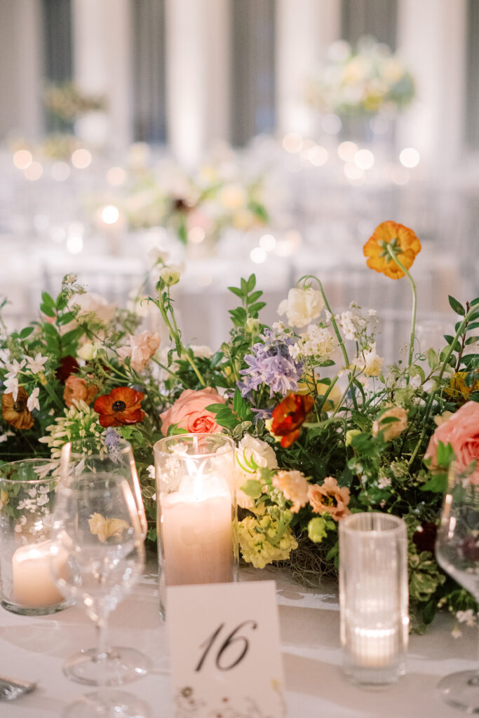 Elegant wedding reception tables feature low lush floral centerpieces and elevated centerpieces in glass vases. The centerpiece is a beautiful arrangement of mixed flowers in soft pastel colors, including yellows, whites, and some darker tones like red and burgundy. Winter wedding reception with floral emphasis and clean lines. Taupe candles in clear glass holders create romantic candlelight. Design by Rosemary & Finch Floral Design. 