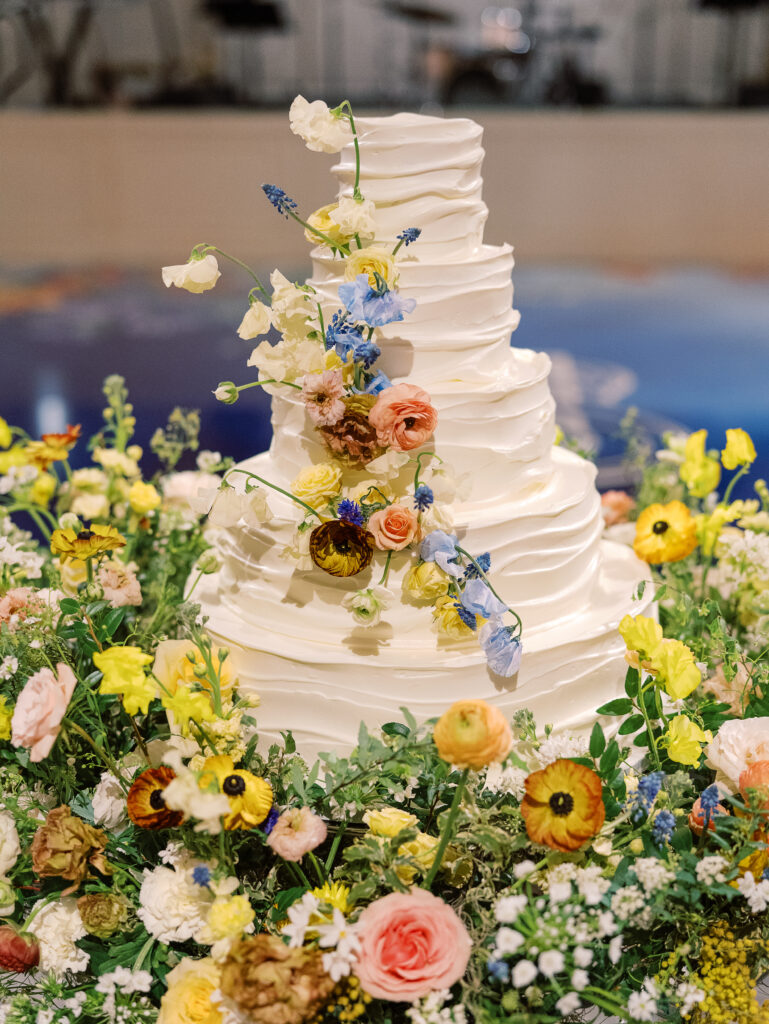 Lush floral cake meadows create memorable moment for wedding reception cake cutting. Elegant winter wedding color palette in reds, yellows, cream, and soft hints of blue. Lush garden-inspired floral installations. Design by Rosemary & Finch Floral Design. 