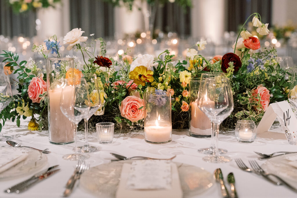 Elegant wedding reception tables feature low lush floral centerpieces and elevated centerpieces in glass vases. The centerpiece is a beautiful arrangement of mixed flowers in soft pastel colors, including yellows, whites, and some darker tones like red and burgundy. Winter wedding reception with floral emphasis and clean lines. Taupe candles in clear glass holders create romantic candlelight. Design by Rosemary & Finch Floral Design. 