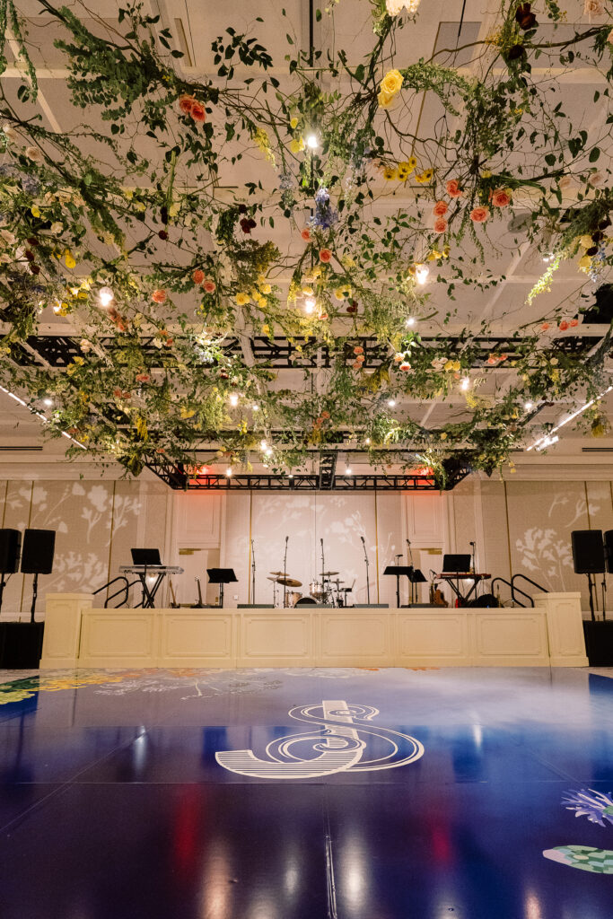 Airy garden-inspired floral installation hanging over wedding reception dance floor. Vining floral garden inspired installation gives interest to the dance floor of this winter wedding in downtown Nashville. Mixed flowers in soft pastel colors, including yellows, whites, and some darker tones like red and burgundy. Design by Rosemary & Finch Floral Design in Nashville, TN.