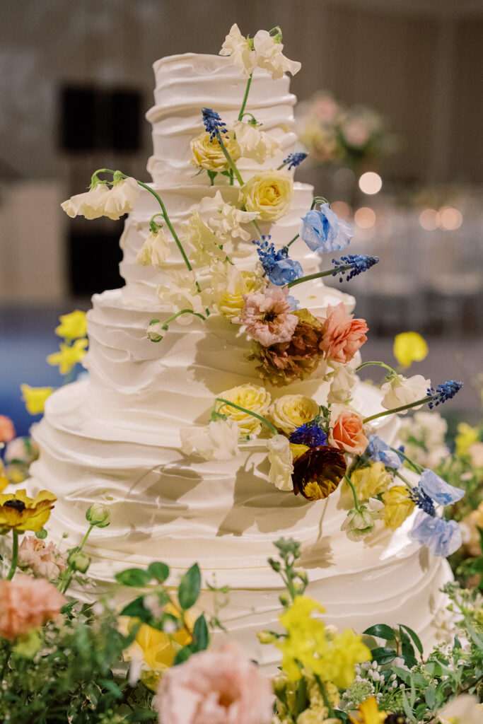 Lush floral cake meadows create memorable moment for wedding reception cake cutting. Elegant winter wedding color palette in reds, yellows, cream, and soft hints of blue. Lush garden-inspired floral installations. Design by Rosemary & Finch Floral Design. 