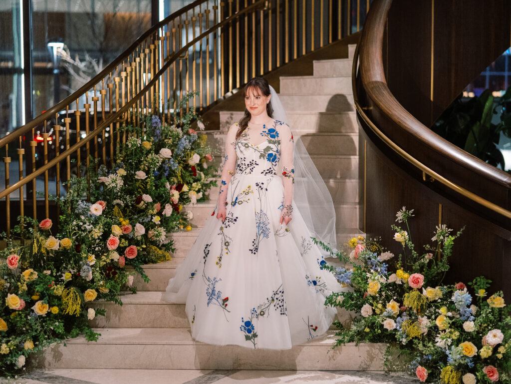 Garden-inspired floral installation on winding staircase. Flowers consisting of soft and vibrant hues, including shades of pink, peach, yellow, cream, and touches of burgundy, adding depth and interest. Winter wedding in downtown Nashville. Unique ceremony entrance for winter wedding. Design by Rosemary & Finch Floral Design. 