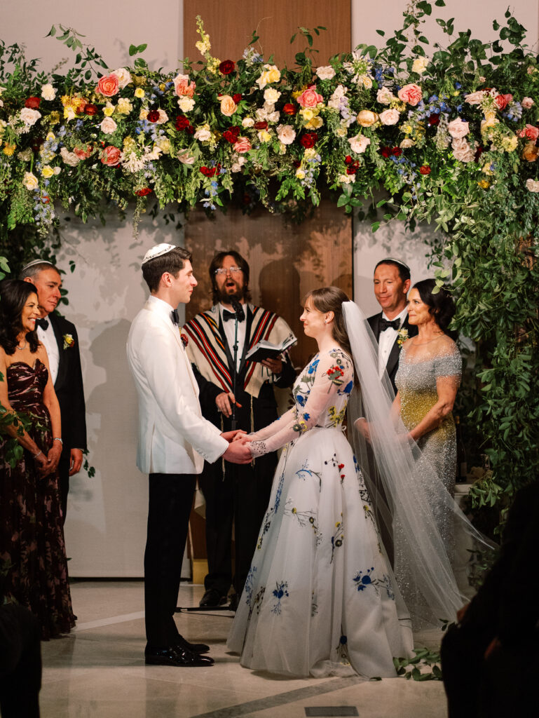 Winter wedding ceremony chuppah wrapped densely in cascading greenery, giving the structure a natural, garden-like appearance. The flowers consisting of soft and vibrant hues, including shades of pink, peach, yellow, cream, and touches of burgundy, adding depth and interest. Winter wedding in downtown Nashville. Design by Rosemary & Finch Floral Design. 