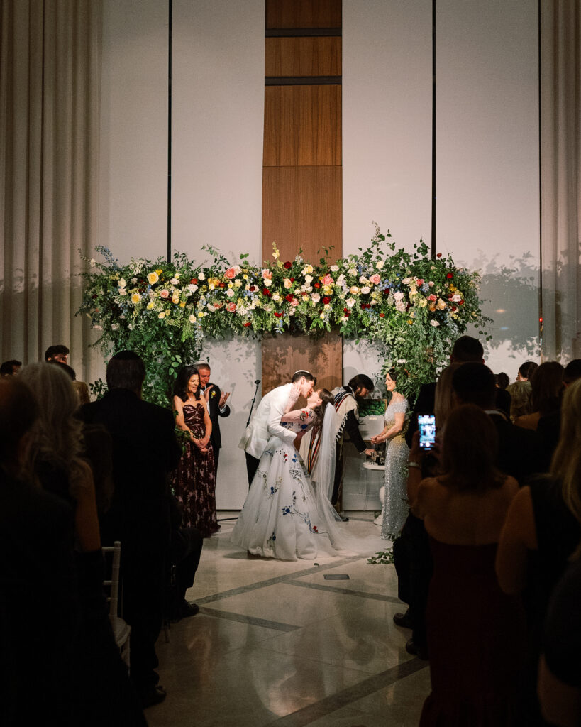 Winter wedding ceremony chuppah wrapped densely in cascading greenery, giving the structure a natural, garden-like appearance. The flowers consisting of soft and vibrant hues, including shades of pink, peach, yellow, cream, and touches of burgundy, adding depth and interest. Winter wedding in downtown Nashville. Design by Rosemary & Finch Floral Design. 