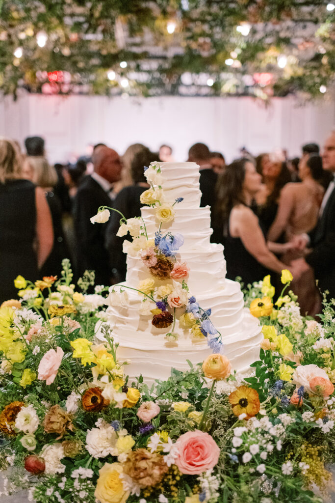 Lush floral cake meadows create memorable moment for wedding reception cake cutting. Elegant winter wedding color palette in reds, yellows, cream, and soft hints of blue. Lush garden-inspired floral installations. Design by Rosemary & Finch Floral Design. 