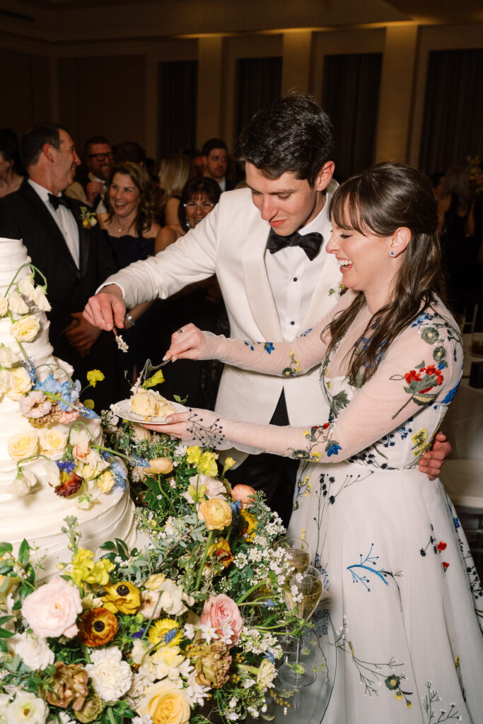 Lush floral cake meadows create memorable moment for wedding reception cake cutting. Elegant winter wedding color palette in reds, yellows, cream, and soft hints of blue. Lush garden-inspired floral installations. Design by Rosemary & Finch Floral Design. 