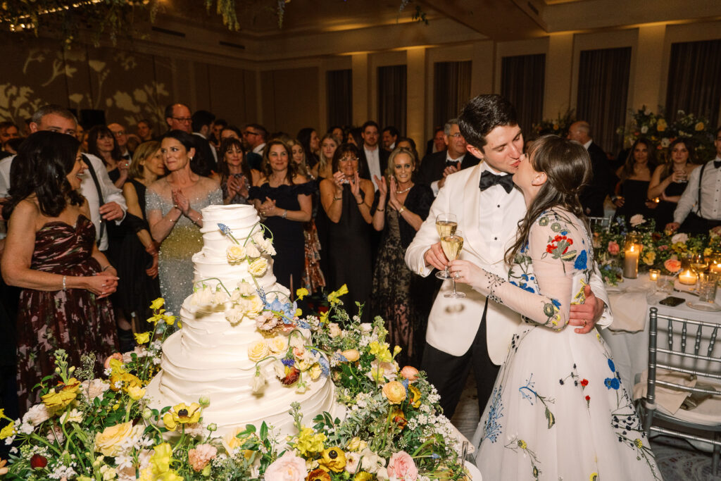 Lush floral cake meadows create memorable moment for wedding reception cake cutting. Elegant winter wedding color palette in reds, yellows, cream, and soft hints of blue. Lush garden-inspired floral installations. Design by Rosemary & Finch Floral Design. 