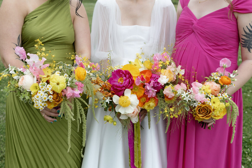 Vibrant and colorful bridal bouquet with flowers that includes lush, fully bloomed peonies in rich magenta, complemented by bright orange and yellow poppies. Summer bridal and bridesmaids’ bouquet with unique and bold spring colors. Whimsical wedding floral with statement making colors. Design by Rosemary & Finch Floral Design. 