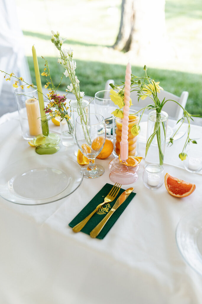 Spring floral centerpieces in bright, cheerful colors, featuring pink peonies, white roses, and wildflowers in orange and yellow. Styled citrus fruits, such as oranges, lemons, limes, and grapefruits for wedding reception tables. Spring wedding with vibrant and unique floral design. Colored glass candle sticks and spiral tapers add unique candle design. Design by Rosemary & Finch Floral Design in Nashville, TN. 