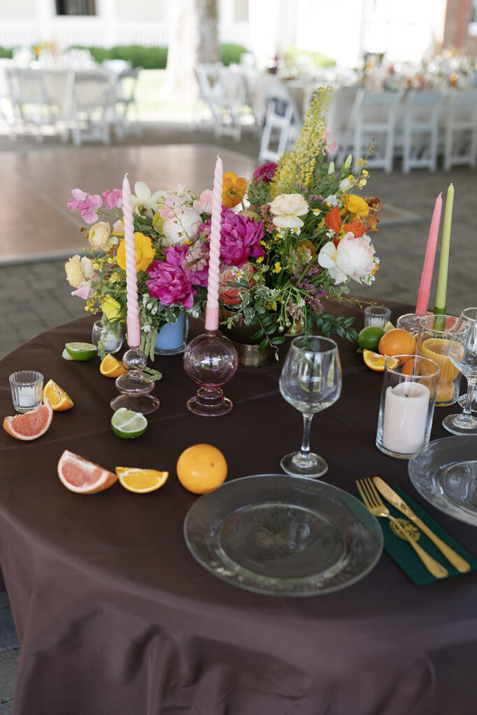 Spring floral centerpieces in bright, cheerful colors, featuring pink peonies, white roses, and wildflowers in orange and yellow. Styled citrus fruits, such as oranges, lemons, limes, and grapefruits for wedding reception tables. Spring wedding with vibrant and unique floral design. Colored glass candle sticks and spiral tapers add unique candle design. Design by Rosemary & Finch Floral Design in Nashville, TN. 