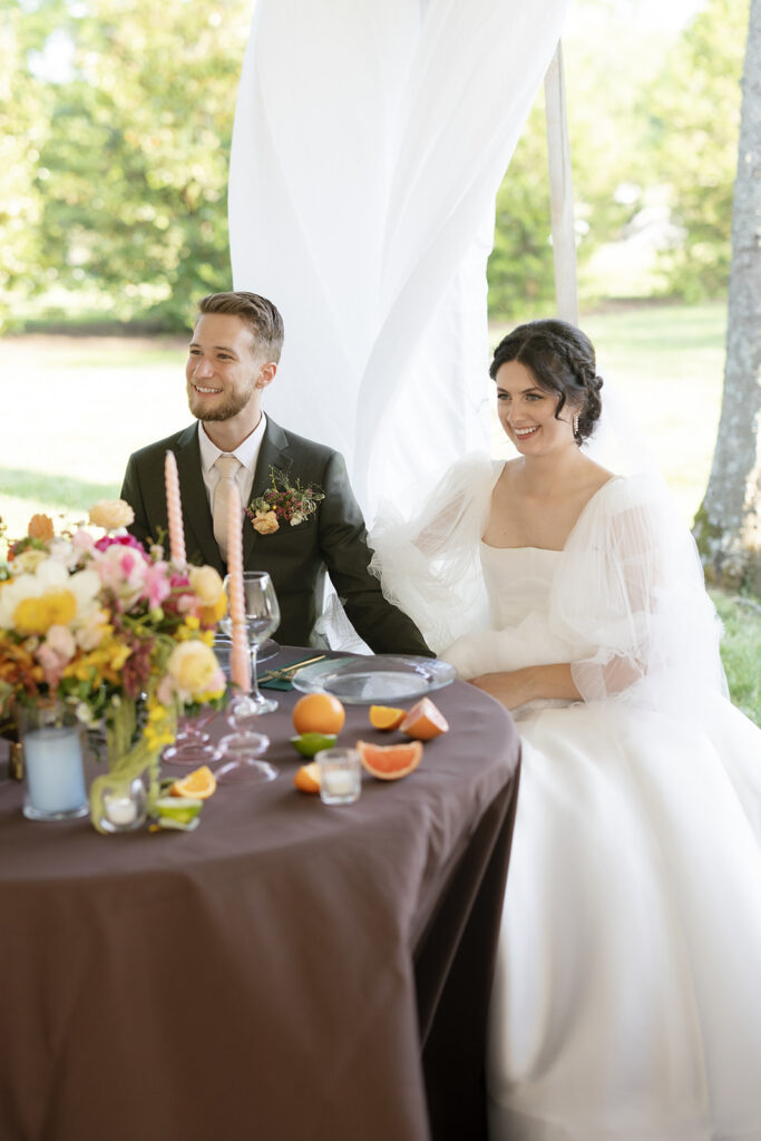 Spring floral centerpieces in bright, cheerful colors, featuring pink peonies, white roses, and wildflowers in orange and yellow. Styled citrus fruits, such as oranges, lemons, limes, and grapefruits for wedding reception tables. Spring wedding with vibrant and unique floral design. Colored glass candle sticks and spiral tapers add unique candle design. Design by Rosemary & Finch Floral Design in Nashville, TN. 