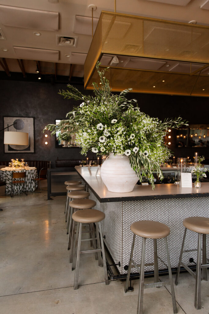 Timeless rehearsal dinner florals in a classic black and white with accents of green color palette. Bud vases and candlelight adorn the tables of this formal rehearsal dinner. Statement urn featuring olive branches and anemones decorate the bar of this reception space. Design by Rosemary & Finch Floral Design in Nashville, TN.