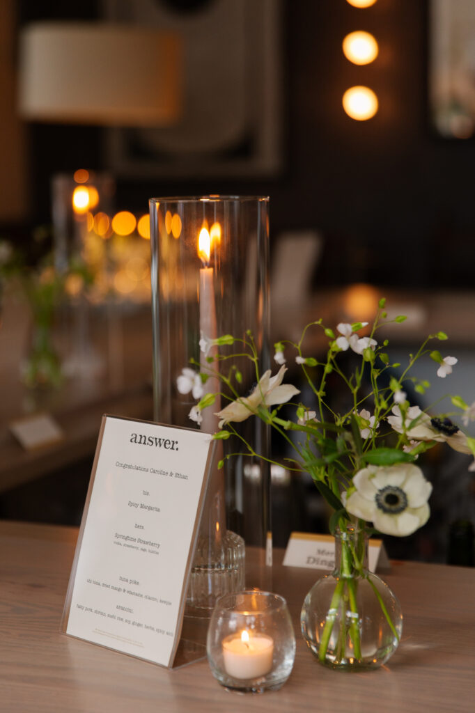 Timeless rehearsal dinner florals in a classic black and white with accents of green color palette. Bud vases and candlelight adorn the tables of this formal rehearsal dinner. Statement urn featuring olive branches and anemones decorate the bar of this reception space. Design by Rosemary & Finch Floral Design in Nashville, TN.